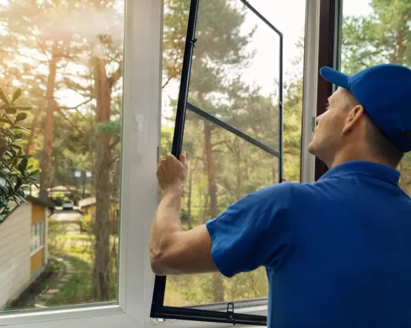 Man in blue shirt and cap performs window screen installation, preparing to fit the mesh into the frame.