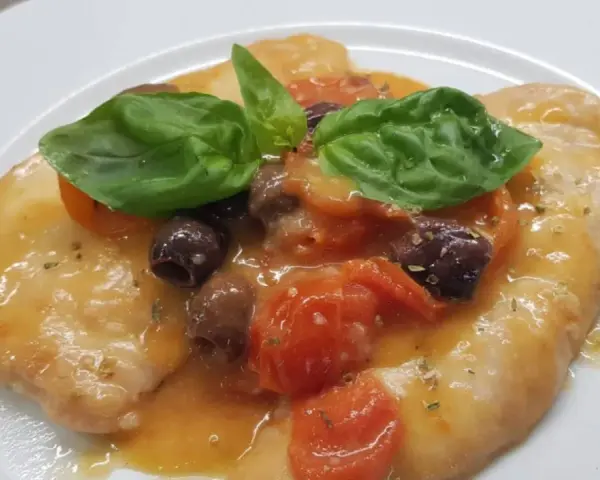 Delizioso pollo alla mediterranea con pomodorini, olive nere e basilico fresco su un piatto bianco.