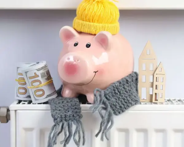 Świnka-skarbonka w czapce i szaliku, z banknotami i domkami na grzejniku. Oszczędzanie na rachunki za ogrzewanie, czyli bon ciepłowniczy w praktyce.