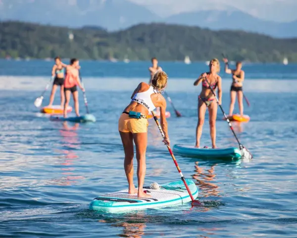 Eine Gruppe von Frauen genießt das Stand-Up-Paddling auf einem See. Sie stehen auf ihren Boards und paddeln durch das ruhige Wasser, während im Hintergrund Berge und Bäume zu sehen sind.