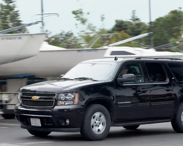 Czarny Chevrolet Suburban jedzie obok przyczep z łodziami, w tle drzewa.