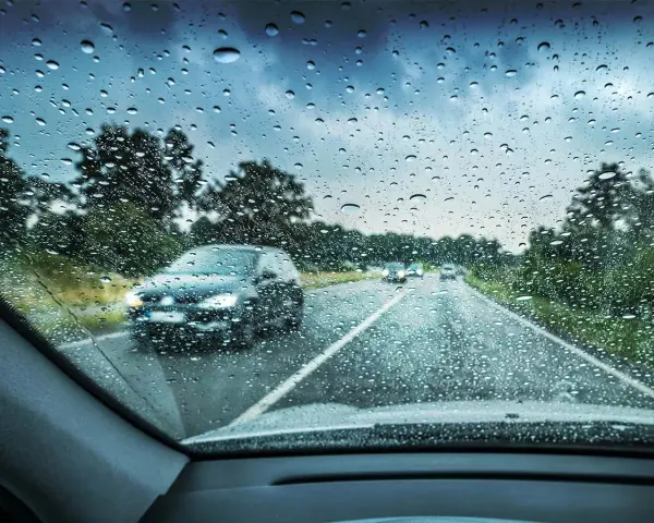 Regndroppar på vindrutan. Bilar kör på en väg genom ett trädbevuxet landskap. Glaco-behandlingen håller sikten klar.