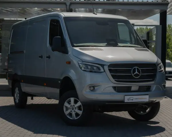 Srebrna budowa ciężarówki, model Mercedesa Sprintera, stoi na parkingu przed nowoczesnym budynkiem.
