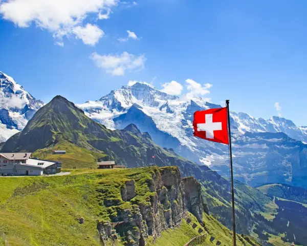A Swiss flag flies proudly against a backdrop of majestic snow-capped mountains and lush green slopes, showcasing the beautiful places of nature in Switzerland.