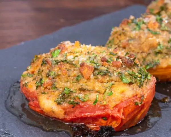 Pomodori gratinati in padella, con croccante panatura alle erbe e formaggio, serviti su una pietra scura.