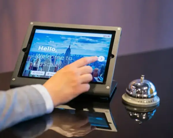 Dłoń dotyka ekranu tabletu z napisem "Welcome to UpScale". Czy można zarezerwować hotel w wieku 17 lat? Obok dzwonek hotelowy.