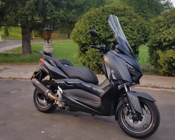 Czarny skuter Yamaha XMAX 300 zaparkowany na tle zieleni i drzew.