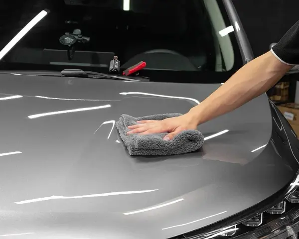 A hand buffs a car's hood with a microfiber cloth, applying a ceramic wax for a deep shine.