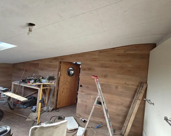 Taket är målat OSB. Rummet är under renovering med träpanel på väggarna, en dörr med runt fönster och byggmaterial utspritt.