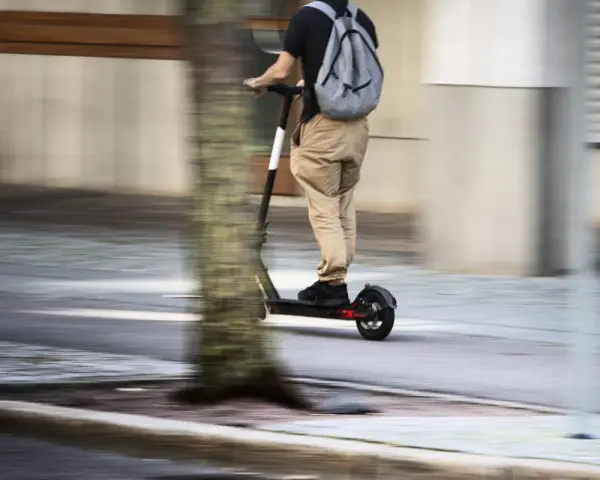 En person på elsparkcykel, med ryggsäck, susar fram på trottoaren. Frågan om får man åka två på elsparkcykel är relevant här.