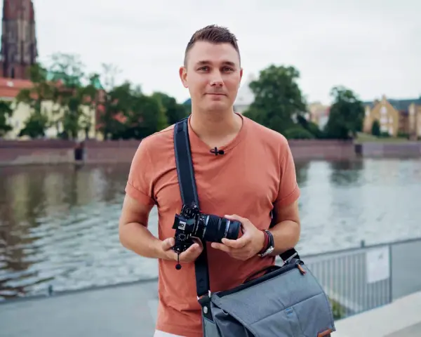 Mężczyzna z aparatem, gotowy na kurs EOS. W tle rzeka i zabytkowe budynki.
