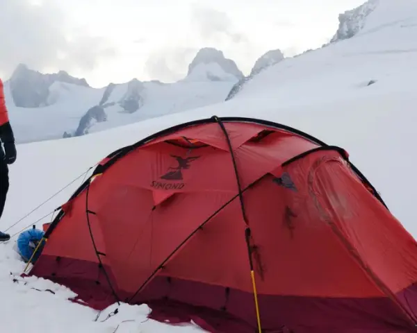 Alpinista w czerwonej kurtce stoi obok namiotu na tle ośnieżonych szczytów. W oddali widać groźne góry, gdzie mogą wystąpić lawiny w Alpach.