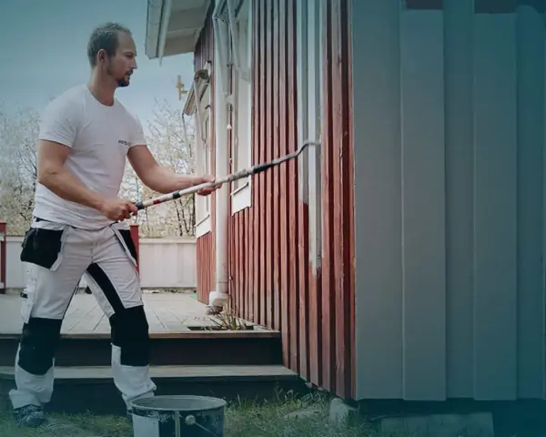 Man i arbetskläder använder en lång roller för att måla huset. En hink med färg står på gräset.