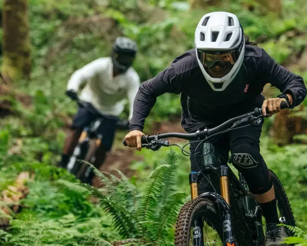 Riders in helmets and protective gear shredding a forest trail. This image shows what to wear mountain biking: full-face helmets, goggles, long sleeves, and knee pads.