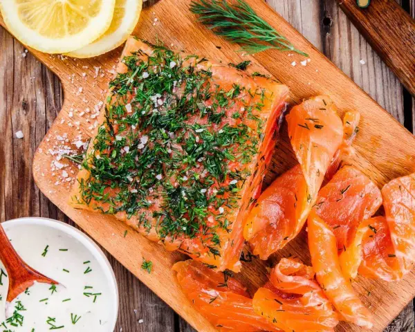En bit gravad lax, generöst toppad med dill och flingsalt, ligger på en träskärbräda bredvid skivor av lax och en skål med sås.