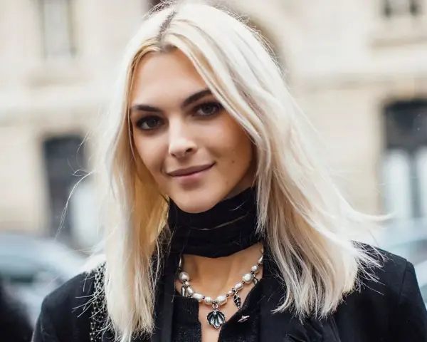 Kobieta z platynowym blondem, w czarnym golfie i naszyjnikiem z pereł, uśmiecha się lekko.