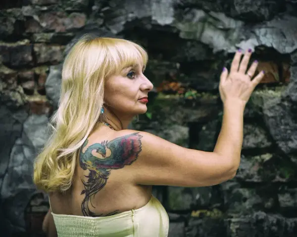 Blondynka z tatuażem feniksa na plecach opiera się o kamienną ścianę. Czyżby moda na tatuaże mijała, a ona wciąż pielęgnuje swoje?