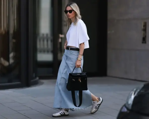 Kobieta w białej bluzce i długiej spódnicy jeansowej, z czarną torebką i sneakersami. Stylowe długa spódnica jeansowa stylizacje.