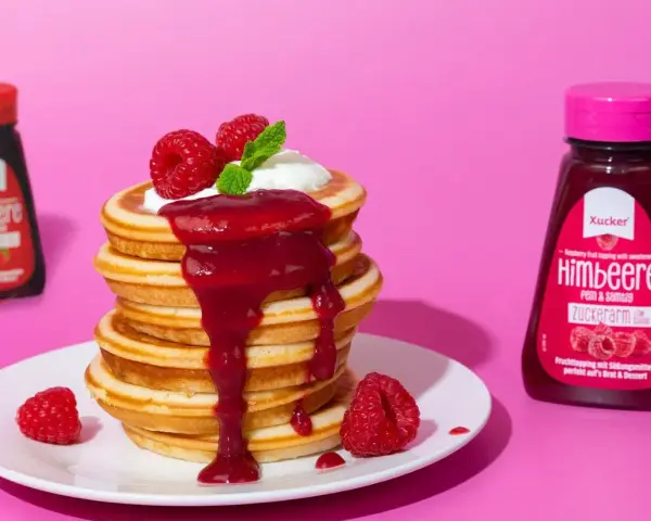 Ein Stapel zuckerfreie pancakes mit Himbeersauce und frischen Beeren. Daneben Flaschen mit Erdbeer- und Himbeer-Topping.