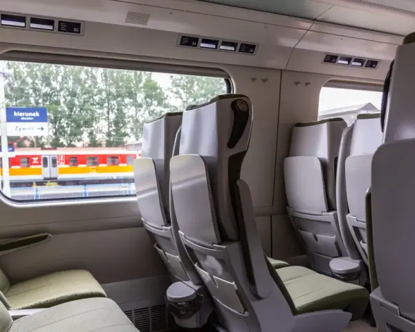 Wnętrze wagonu bez przedziałów IC. Rozkład miejsc z zielonymi siedzeniami i stolikami. Za oknem peron i pociąg.