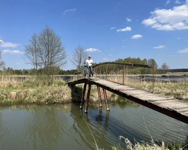 Osoba z rowerem na drewnianym mostku nad rzeką. Piękny dzień na odkrywanie tras rowerowych łódzkiego.