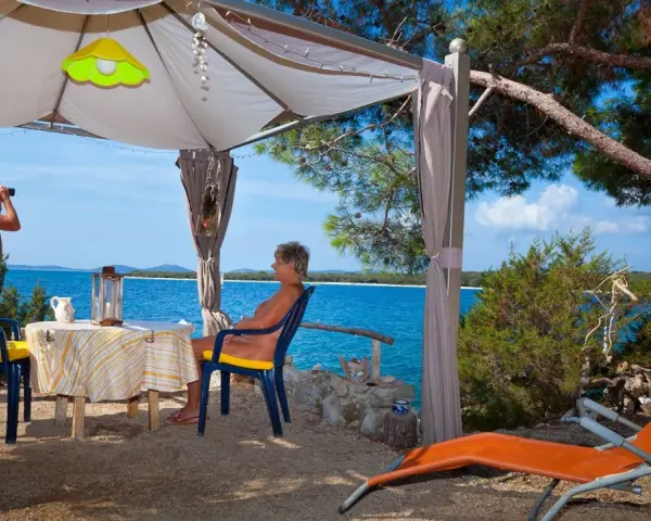 Para na plaży, relaksująca się w namiocie. Mężczyzna obserwuje morze przez lornetkę. To idealne miejsce na naturystyczny camping.