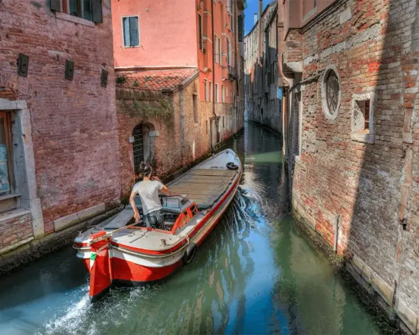 Un uomo guida una barca su un canale stretto a Venezia, circondato da edifici in mattoni. Quanti canali ci sono a Venezia? Molti, come questo!