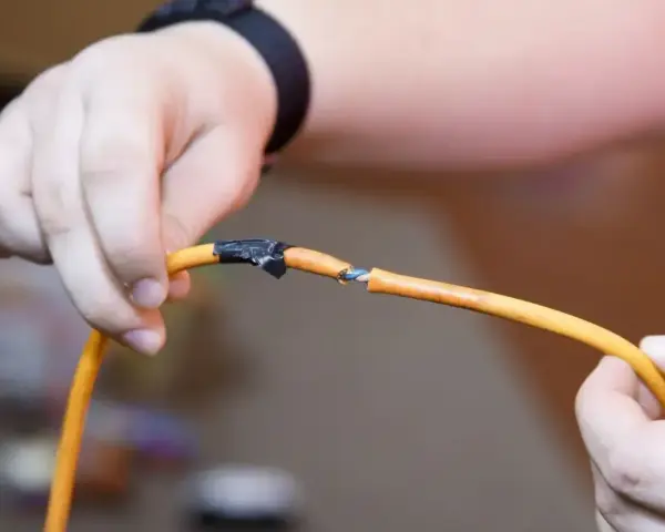 Dłonie trzymające pomarańczowy kabel z odsłoniętymi żyłkami, prowizorycznie połączone taśmą. Pokazuje, jak połączyć kable bez lutowania.