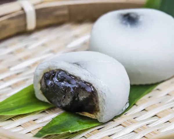 Dwa białe mochi, jedno przekrojone, ukazujące ciemne nadzienie. Podane na bambusowej tacce z liśćmi.