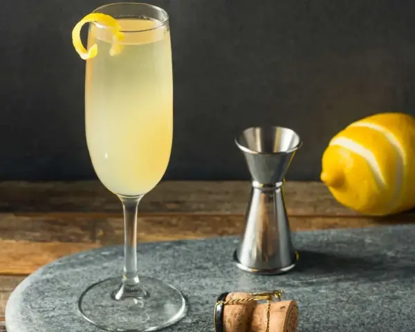 Elegancki drink z szampanem, ozdobiony skórką cytryny, obok kieliszka stoi miarka barmańska, cytryna i korek od szampana.