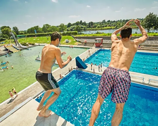 Zwei Jungen springen vom Sprungbrett in den Badesee Bremen. Im Hintergrund sind Wasserrutschen und viele Badegäste zu sehen.
