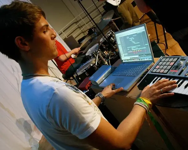 Junger Mann am Laptop und Keyboard, der Musik produziert. Ein Schlagzeuger im Hintergrund. Perfekt für ein Musikproduktion Studium.
