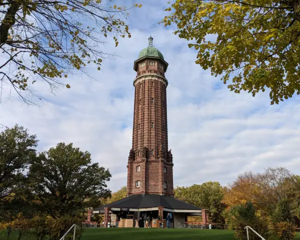 Der Aussichtsturm im Volkspark Jungfernheide ragt mit seinem roten Backstein und grünen Kuppeldach in den Himmel. Herbstlaub umrahmt die Szene.