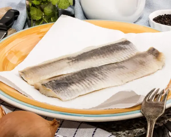 Zwei Matjesfilets auf Küchenpapier, bereit für eine gesunde Mahlzeit. Zwiebeln und Pfefferkörner deuten auf die Zubereitung hin.