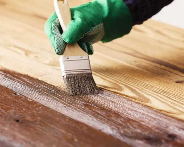 Mano con guante verde aplicando barniz a madera. El proceso de barnizar madera protege y embellece la superficie.