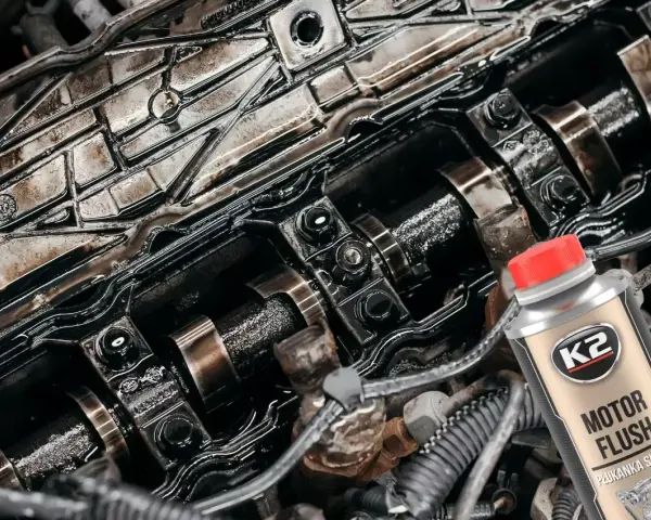Silnik z widocznymi elementami rozrządu, pokryty czarnym osadem. Obok butelka płukanki K2 Motor Flush, która pomoże rozwiązać problem niskie ciśnienie oleju na rozgrzanym silniku.