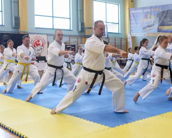 Grupa osób w kimonach ćwiczy karate na niebiesko-żółtej macie w sali gimnastycznej.