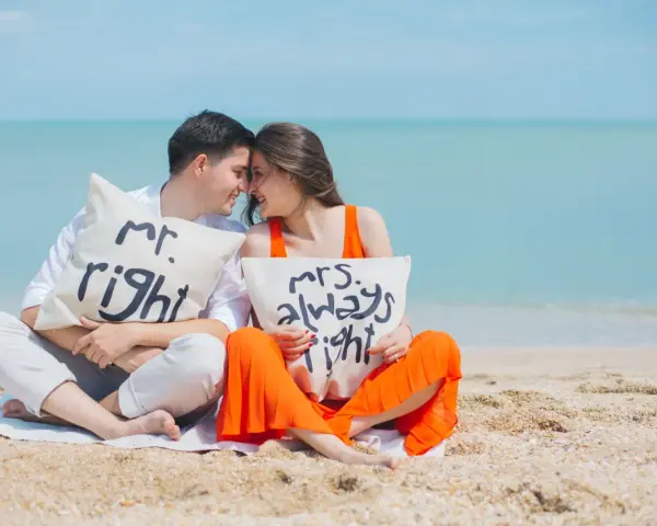Para zakochanych na plaży z poduszkami "Mr. Right" i "Mrs. Always Right". Ich uśmiechy i bliskość symbolizują idealny **kompromis w związku**.