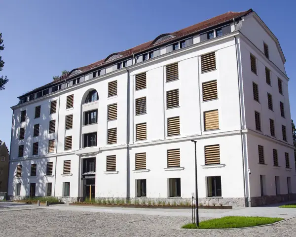 Nowoczesny, biały spichlerz w Słupsku z licznymi oknami o drewnianych żaluzjach, stojący na brukowanym placu.