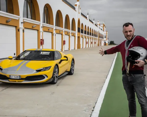 Żółte Ferrari 296 GTB z szarymi pasami na torze wyścigowym. Obok stoi mężczyzna z kaskiem, zapraszając do przejażdżki.