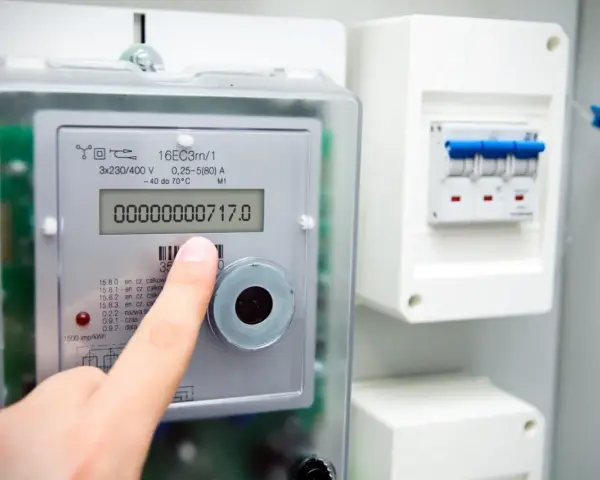Palec wskazuje na cyfrowy licznik energii elektrycznej, pokazujący odczyt 00000000717.0. Widoczne są symbole na liczniku energii elektrycznej.