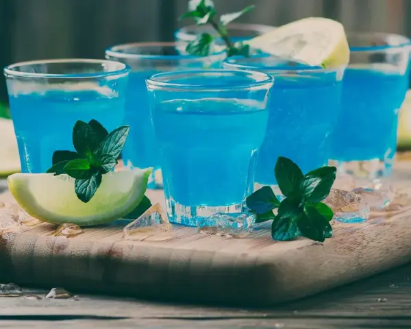 Kilka shotów z drinkiem blue curacao, ozdobionych limonką i miętą, na drewnianej desce z lodem.