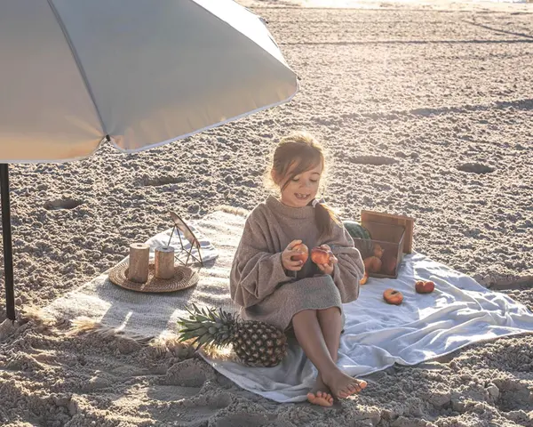 Dziewczynka na plaży pod parasolem, jedząca owoce. Idealny zestaw na wakacje nad morze z dzieckiem: koc, owoce, ananas.