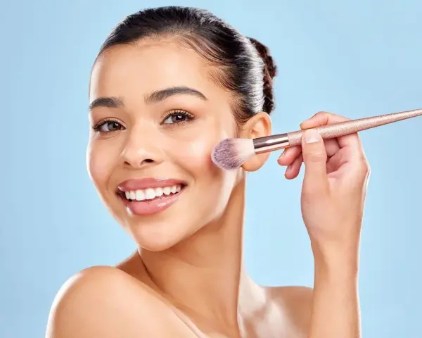 Une femme souriante utilise un pinceau pour appliquer du correcteur maquillage sur sa joue.