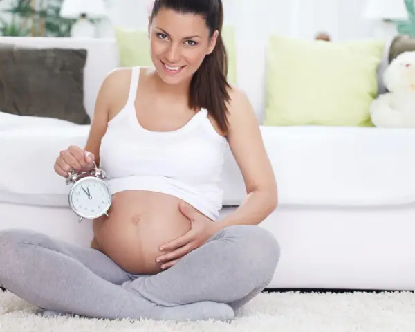 Uśmiechnięta kobieta w 40 tygodniu ciąży trzyma budzik. Czas na poród!