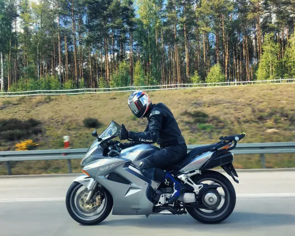 Motocyklista w kasku jedzie na srebrnym Honda VFR 800 wzdłuż leśnej drogi.