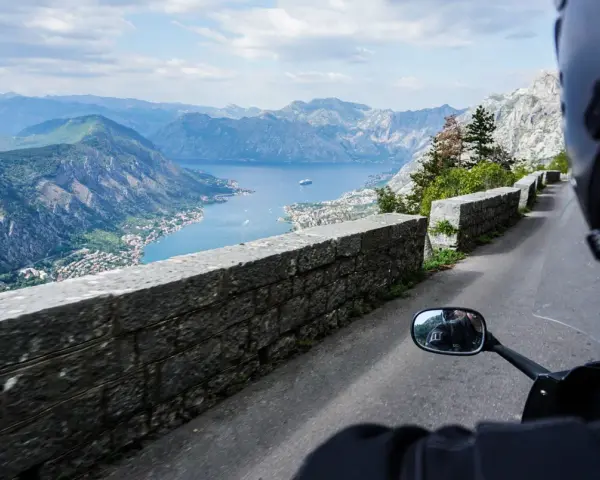 Widok z motocykla na Zatokę Kotorską w Czarnogórze. Kręta droga z kamiennym murem i zapierająca dech w piersiach panorama gór i morza.