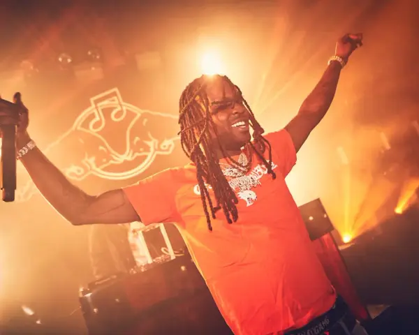 Rapper mit Dreadlocks und orangefarbenem T-Shirt singt mit erhobenen Armen und Mikrofon. Die Bühne ist in warmes Licht getaucht, perfekt für energiegeladene Hip-Hop-Songs.