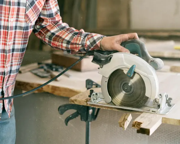 A person in a plaid shirt demonstrates how to use a circular saw, cutting wood with sawdust flying.