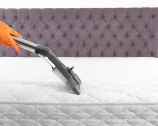 Main tenant, gant orange, utilise un aspirateur pour comment nettoyer un matelas à sec. Le matelas est blanc, avec un couvre-lit capitonné gris en arrière-plan.